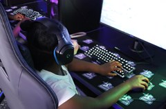 A little girl with her back to the camera sits in a gaming chair, a little too big for her. She looks at the bright computer screen with one hand on a mouse and the other on the keyboard.