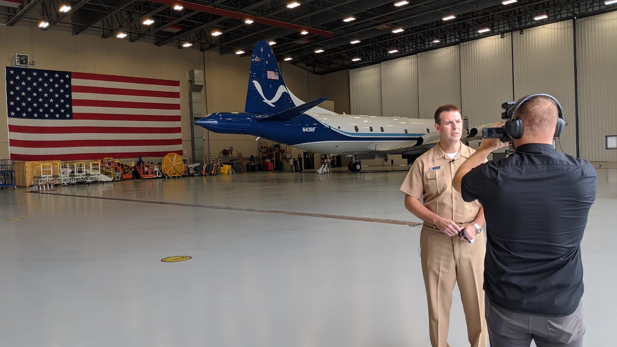 Pilot Sloane being interviewed at the NOAA Hurricane Hunters Hangar at Lakeland Linder Int'l Airport with plane and American flag in background