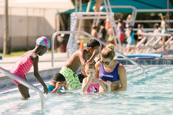 group of children in swim lessons on the steps of Gandy Pool. Little girl in foreground holding nose while working with instructor.