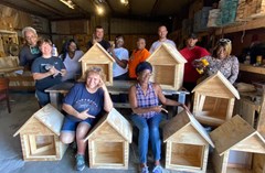 A group photo of 11 neighborhood leaders and workshop instructors gathered around their finished Little Libraries. They are inside a workshop with natural lighting coming in.