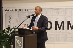 Man in dark suit speaking into a microphone at a podium with poster of Lakeland History & Culture Center swan logo and large map mural in background. Large green plant located to the left of the podium