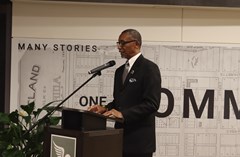 Man in black suit speaking into a microphone at a podium with poster of Lakeland History & Culture Center swan logo and large map mural in background. Large green plant located to the left of the podium