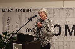 Woman speaking into a microphone at a podium with poster of Lakeland History & Culture Center swan logo and large map mural in background. Large green plant located to the left of the podium