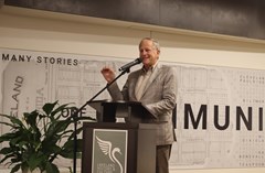 Man in light suit speaking into a microphone at a podium with poster of Lakeland History & Culture Center swan logo and large map mural in background. Large green plant located to the left of the podium