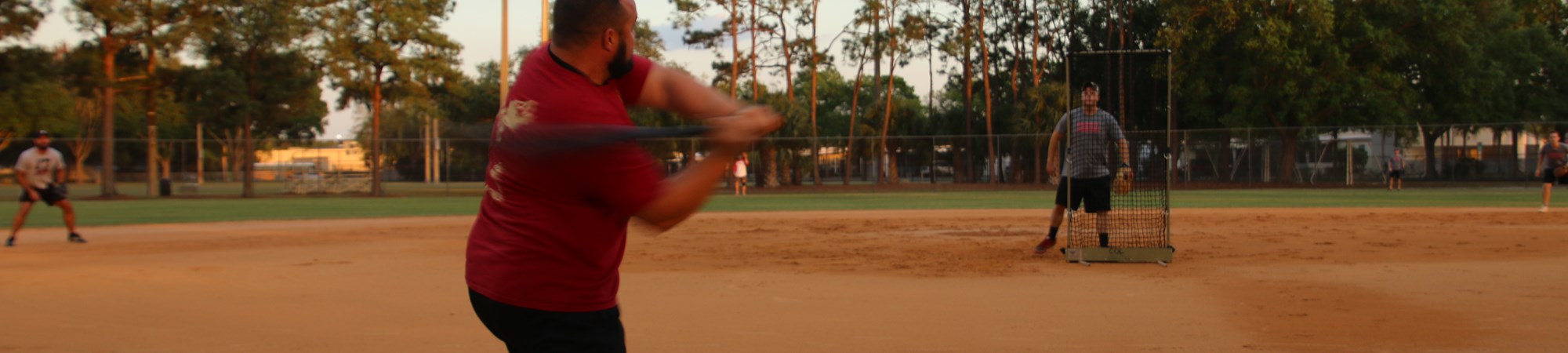Softball field at Sunset, man in the foreground swinging a baseball bat