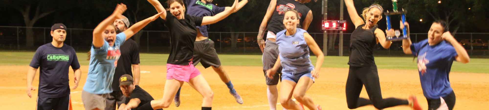 players on a softball field after dark playing kickball. In the foreground on man, wearing neon green shorts and pink shirt is sliding into home while another man lunges to tag him out.