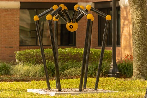 A black long-legged spider with a spherical yellow body, yellow leg joints, and a black circular eye located in the center of the body.