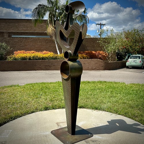 Metal sculpture shaped in an abstract shape of a woman in the foreground titled "eve," brick wall and flowers in the background.