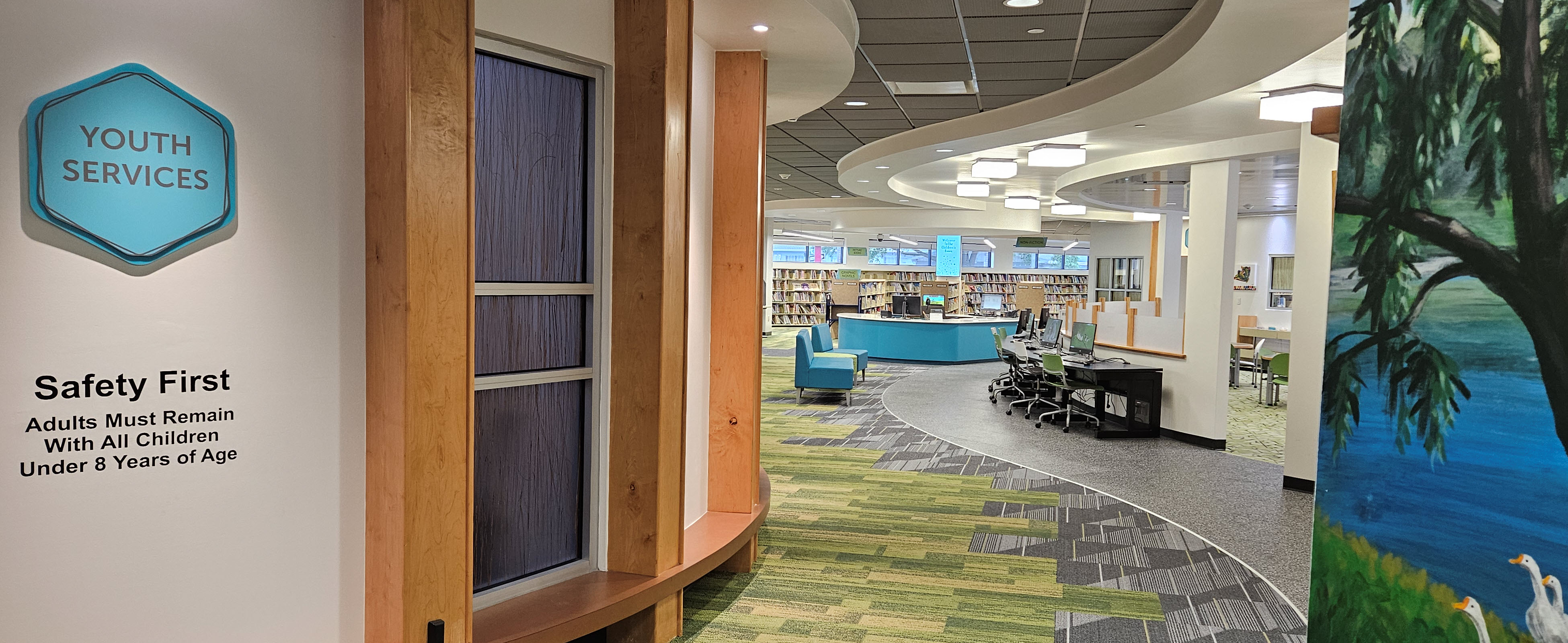 Entrance to Youth Services room with mural wall in foreground, service desk, chairs, computers, and bookshelves in background