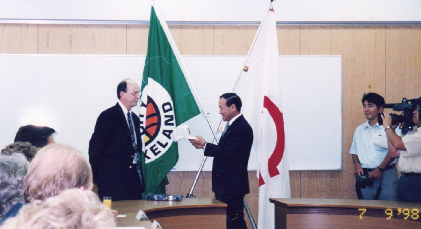 Representatives from the City of Lakeland, Florida and Imabari, Japan meet in Imabari with the flags of two cities in the background to create a friendship contract as part of Sister Cities International, with a man representing Imabari holding a paper in front of a man representing the City of Lakeland with an entourage of Lakelanders and cameramen watching; link to contentDM Sister Cities International collection