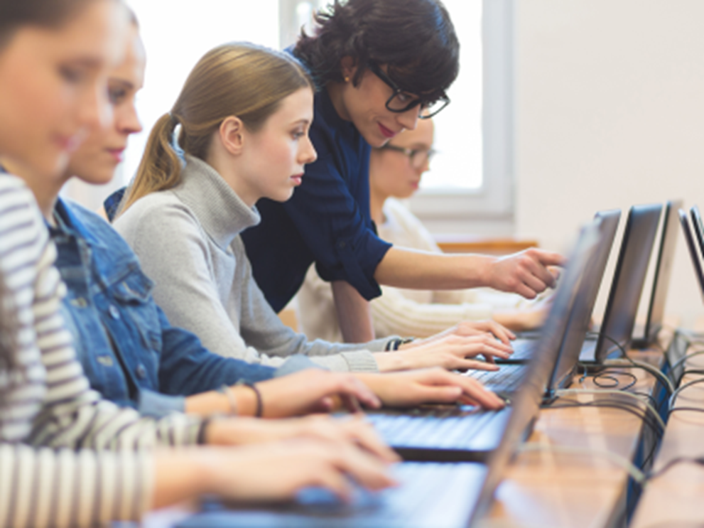 Row of people using laptops at a shared desk with instructor helping one person on their laptop